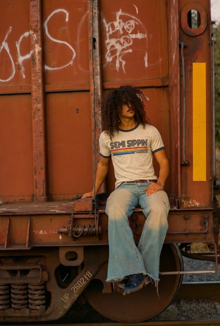 A man in the Nashville ringer sitting on the coupling of a rusted freight car, late sun