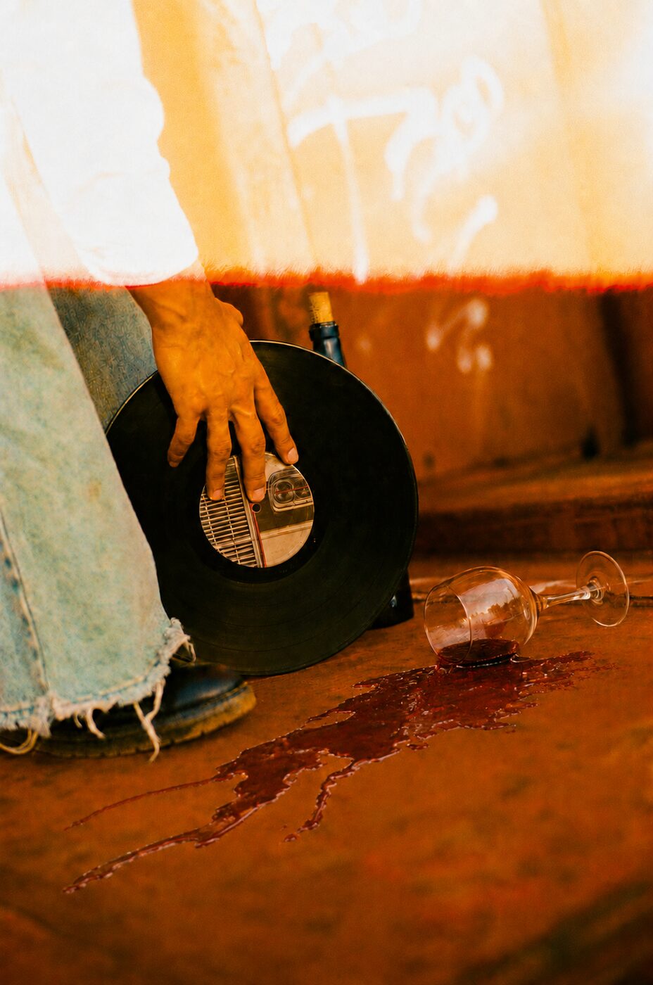 A hand on a vinyl record, red wine spilled across rust-coloured ground
